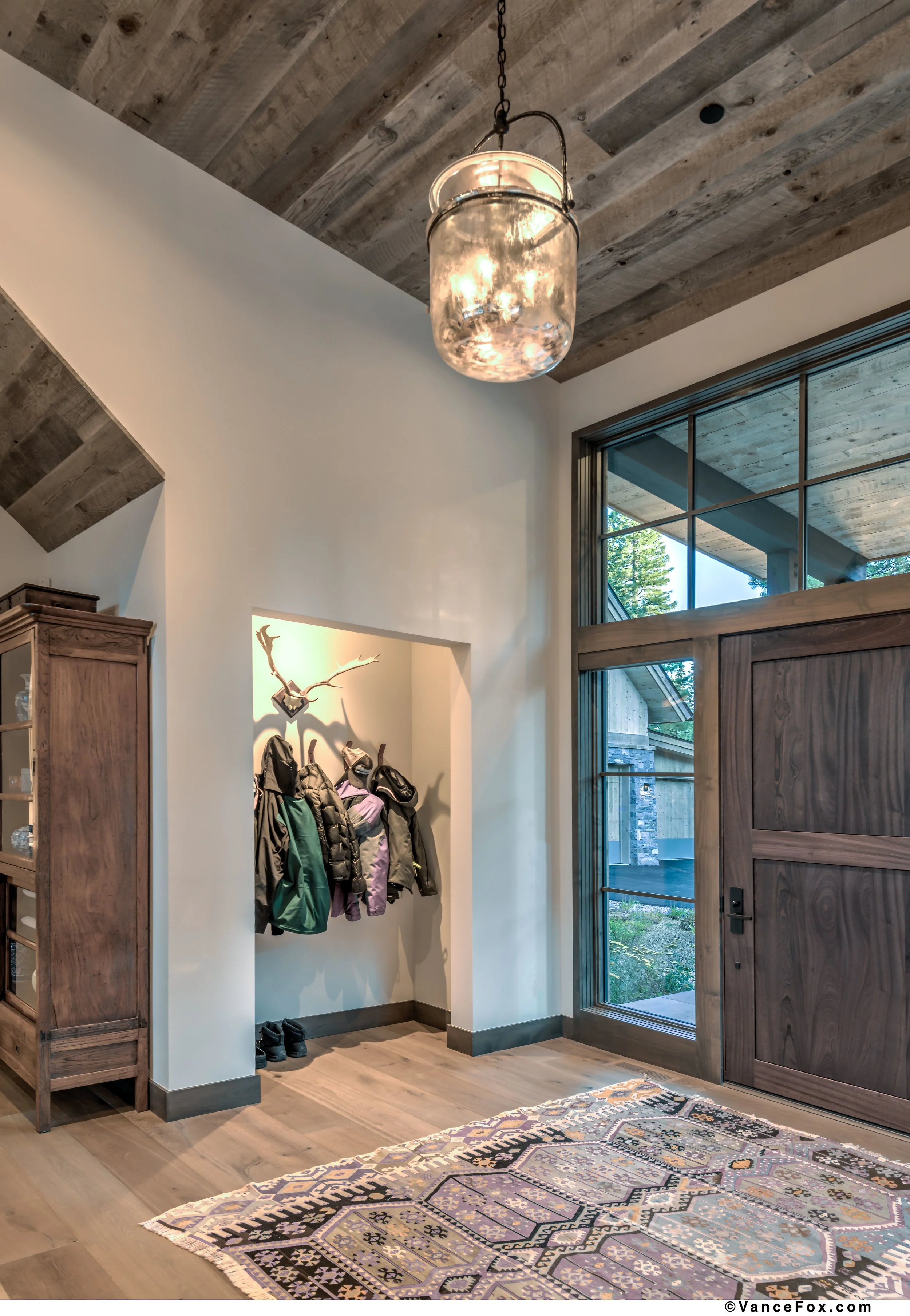 Entryway with wooden door, coat hooks with jackets, shoes on the floor, large window, decorative hanging light, and a colorful area rug.