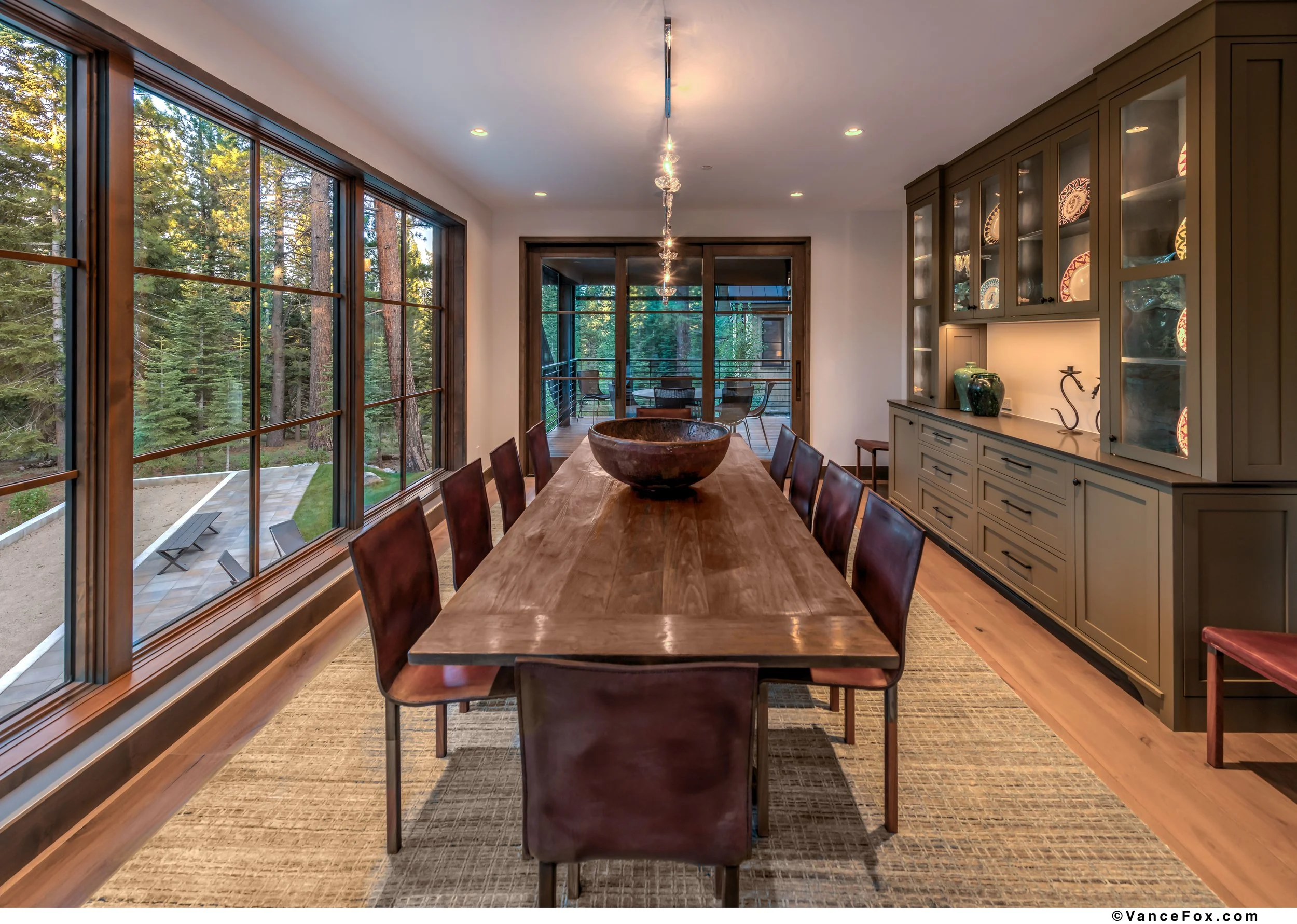 A modern dining room with a long wooden table surrounded by eight leather chairs, large windows showing a forest view, a sideboard with decorative items, and a sliding glass door leading to an outdoor balcony.