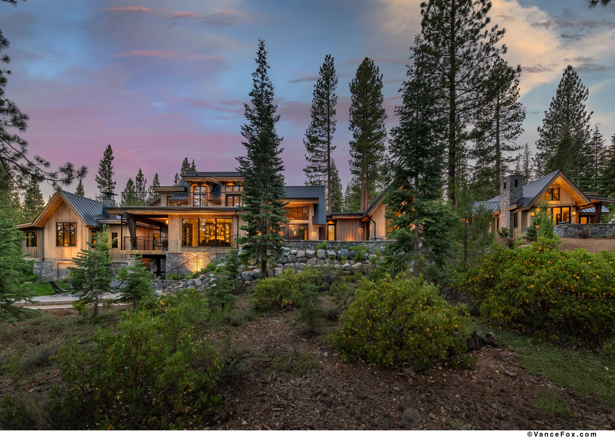 Luxurious house among tall pine trees at dusk, with interior lights glowing through large windows, on a hillside with greenery and rocks.
