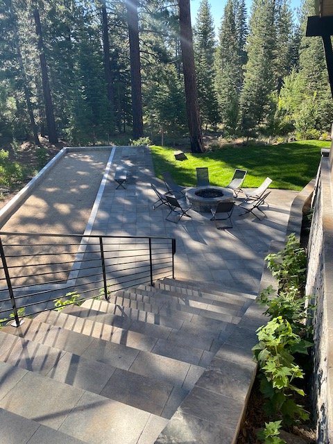 Outdoor patio with stairs, a fire pit surrounded by chairs, and a grassy area with trees in the background.