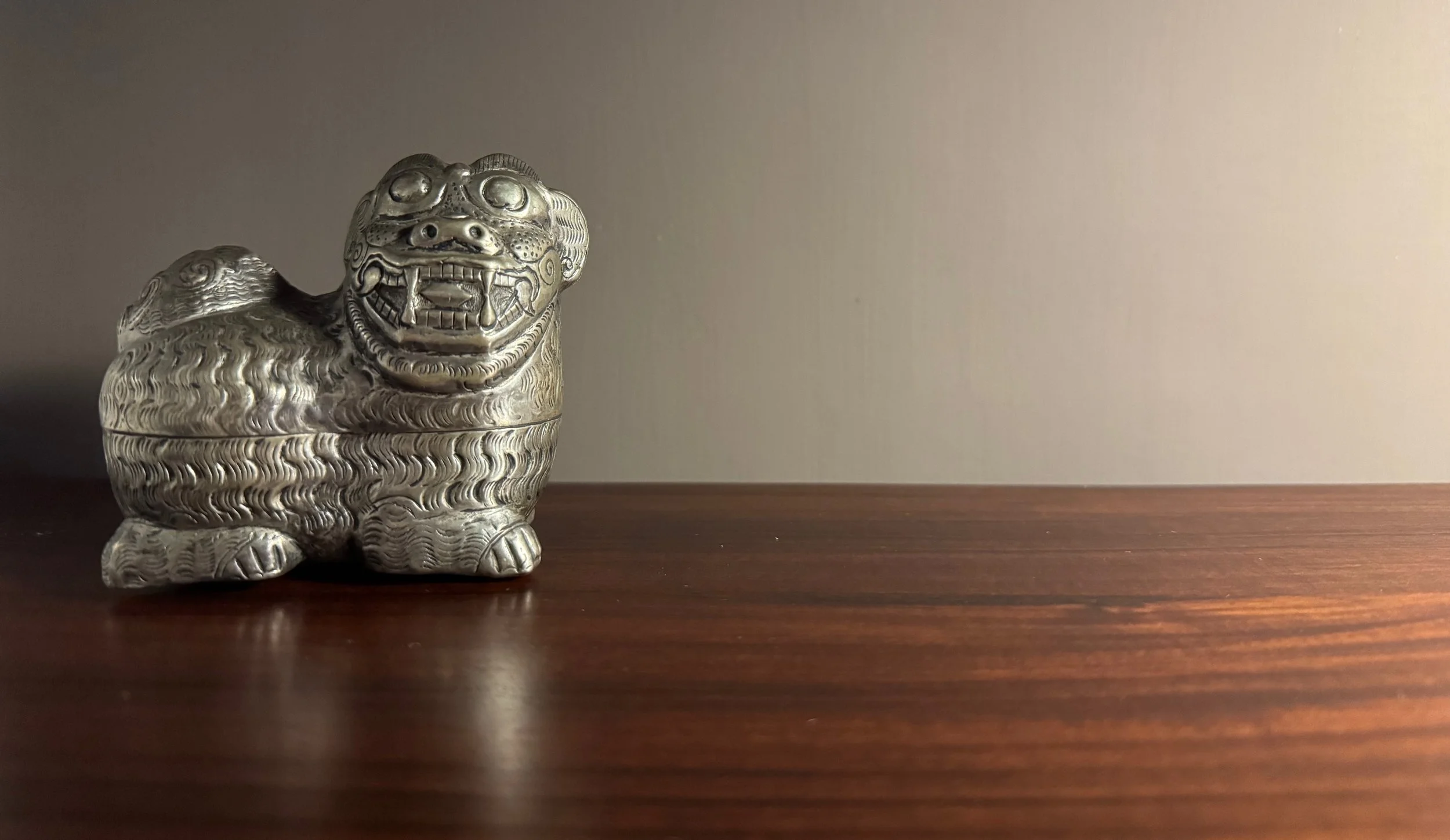 A silver, intricately designed Chinese foo dog or guardian lion figurine on a polished wooden surface against a plain wall.