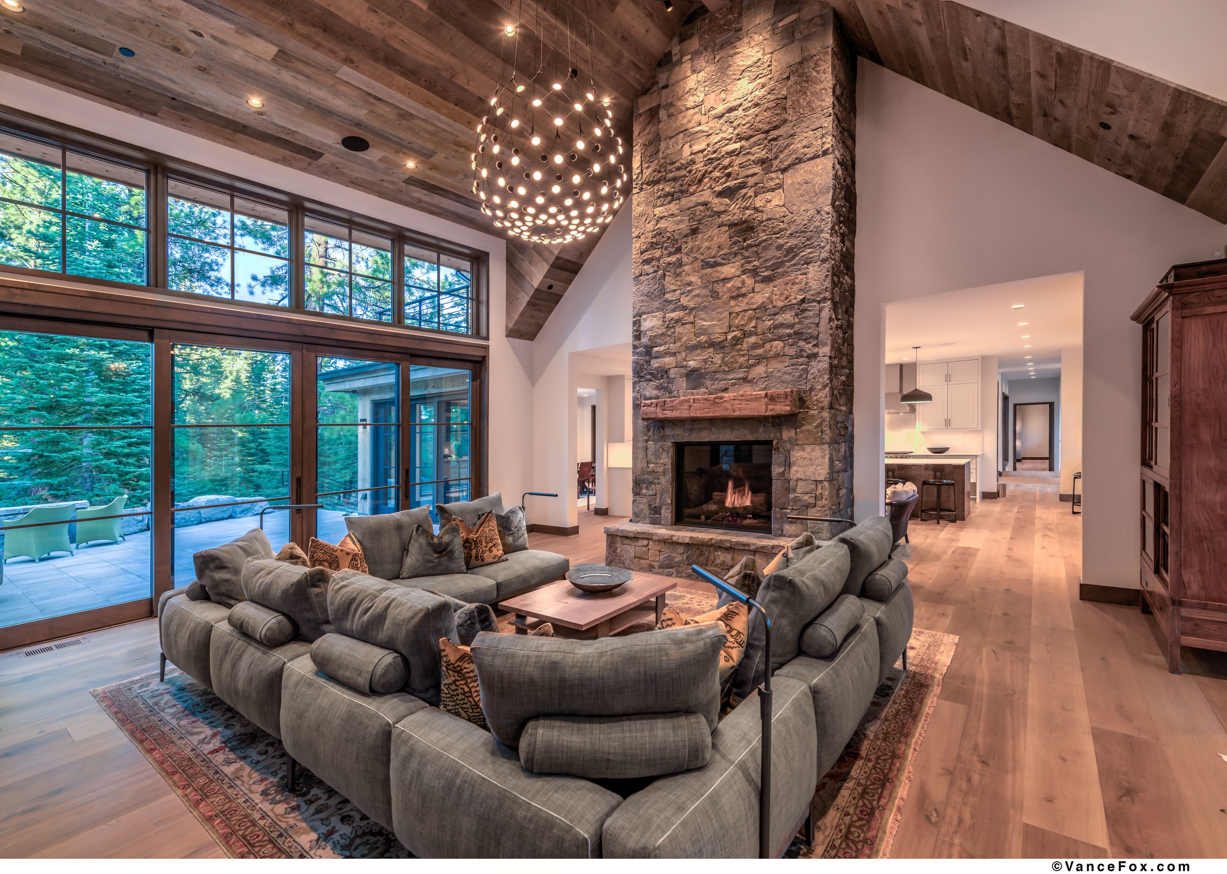 Living room with large windows, a stone fireplace, a round chandelier, and a sectional sofa with decorative pillows.