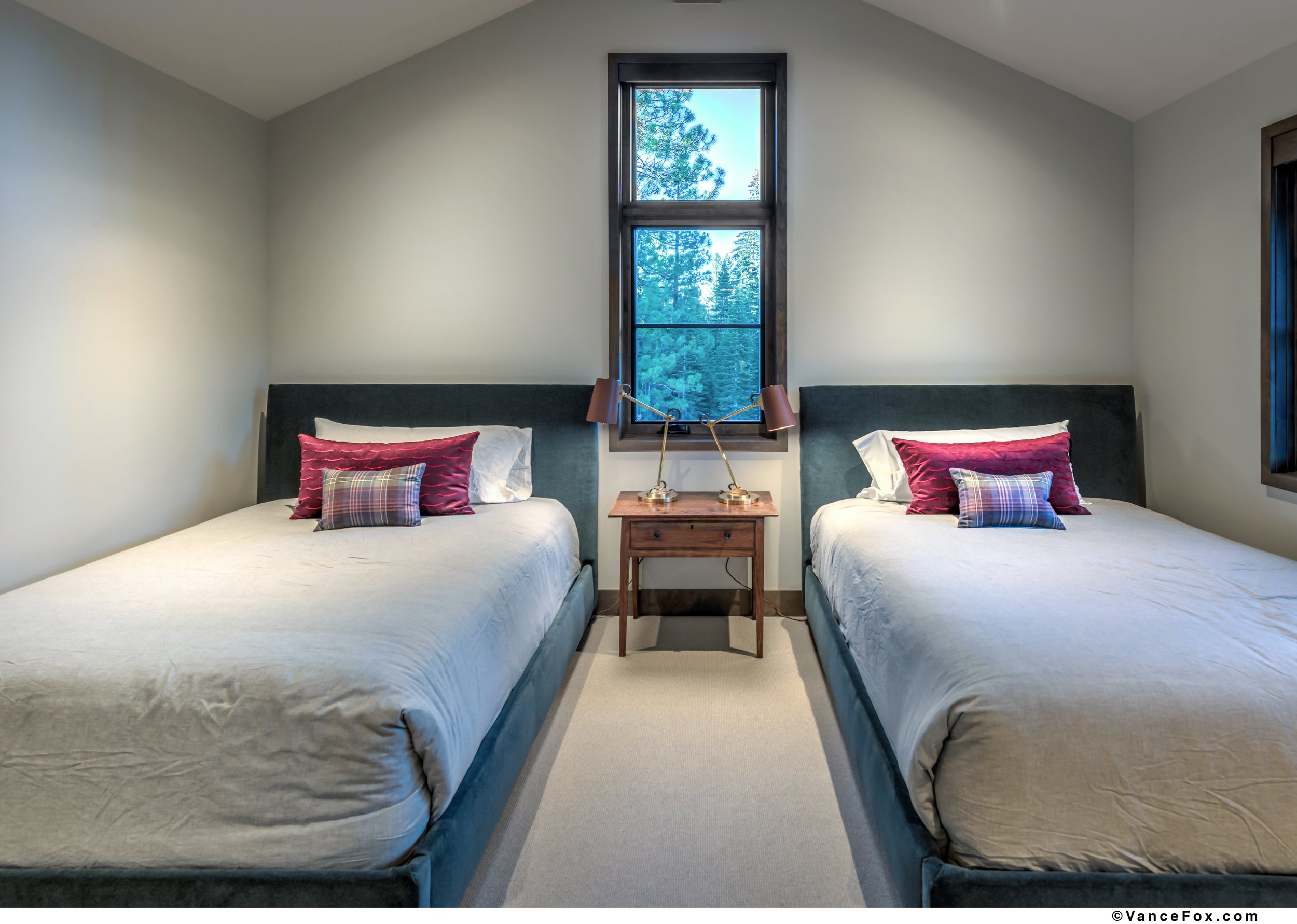 A bedroom with two beds, each with decorative pillows, a wooden nightstand between them, and a window showing trees outside.