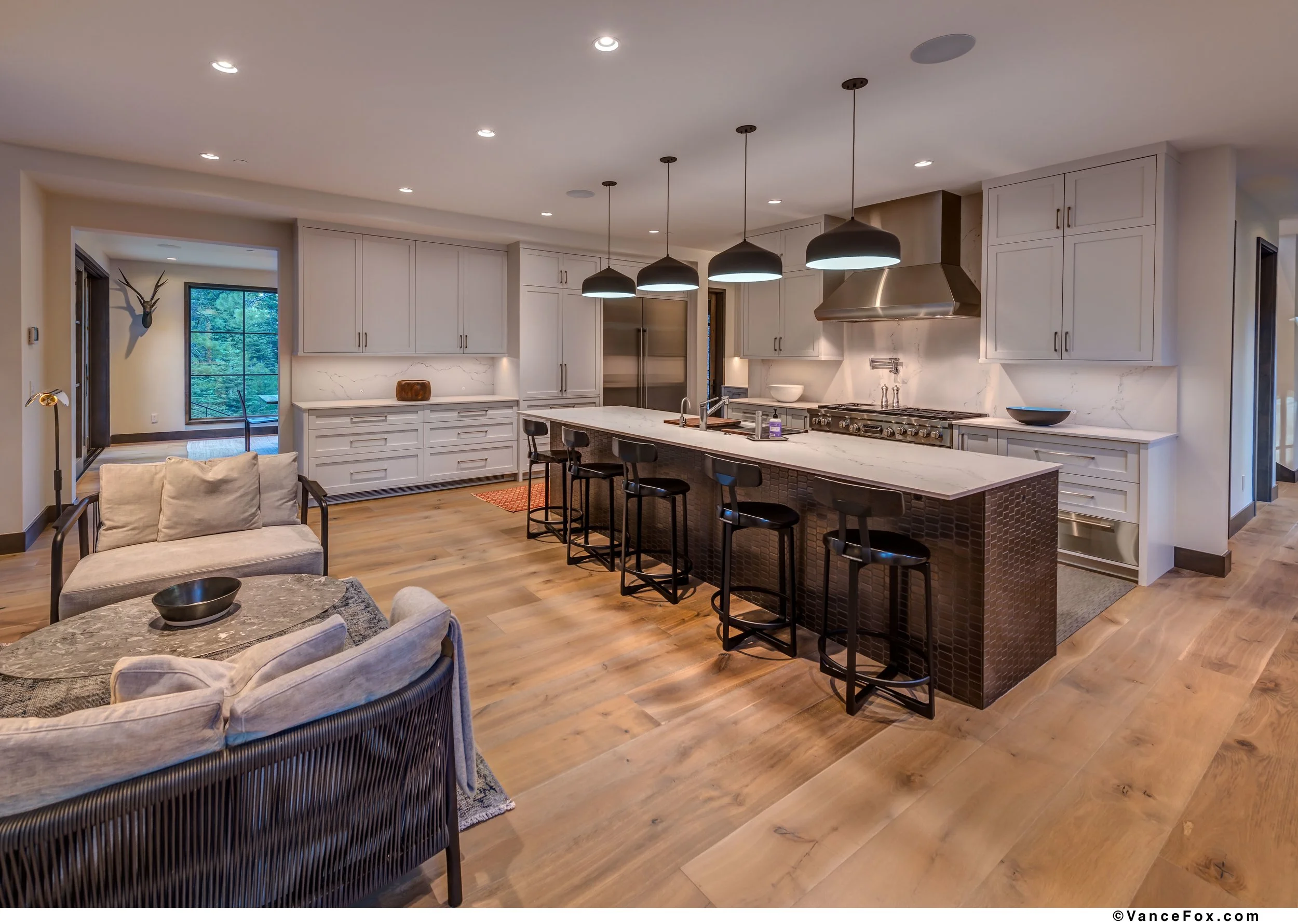 Modern kitchen with white cabinets, marble countertops, and a large island with four black barstools. Pendant lights hang above the island, and there is a stainless steel refrigerator and a stovetop with a vent hood. Adjacent to the kitchen is a seat
