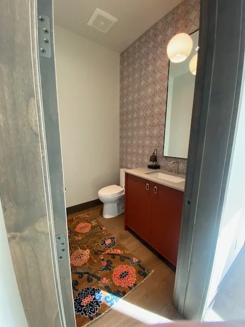 Small bathroom with a toilet, a wooden vanity with a sink, a mirror, patterned wallpaper, and two hanging light fixtures.