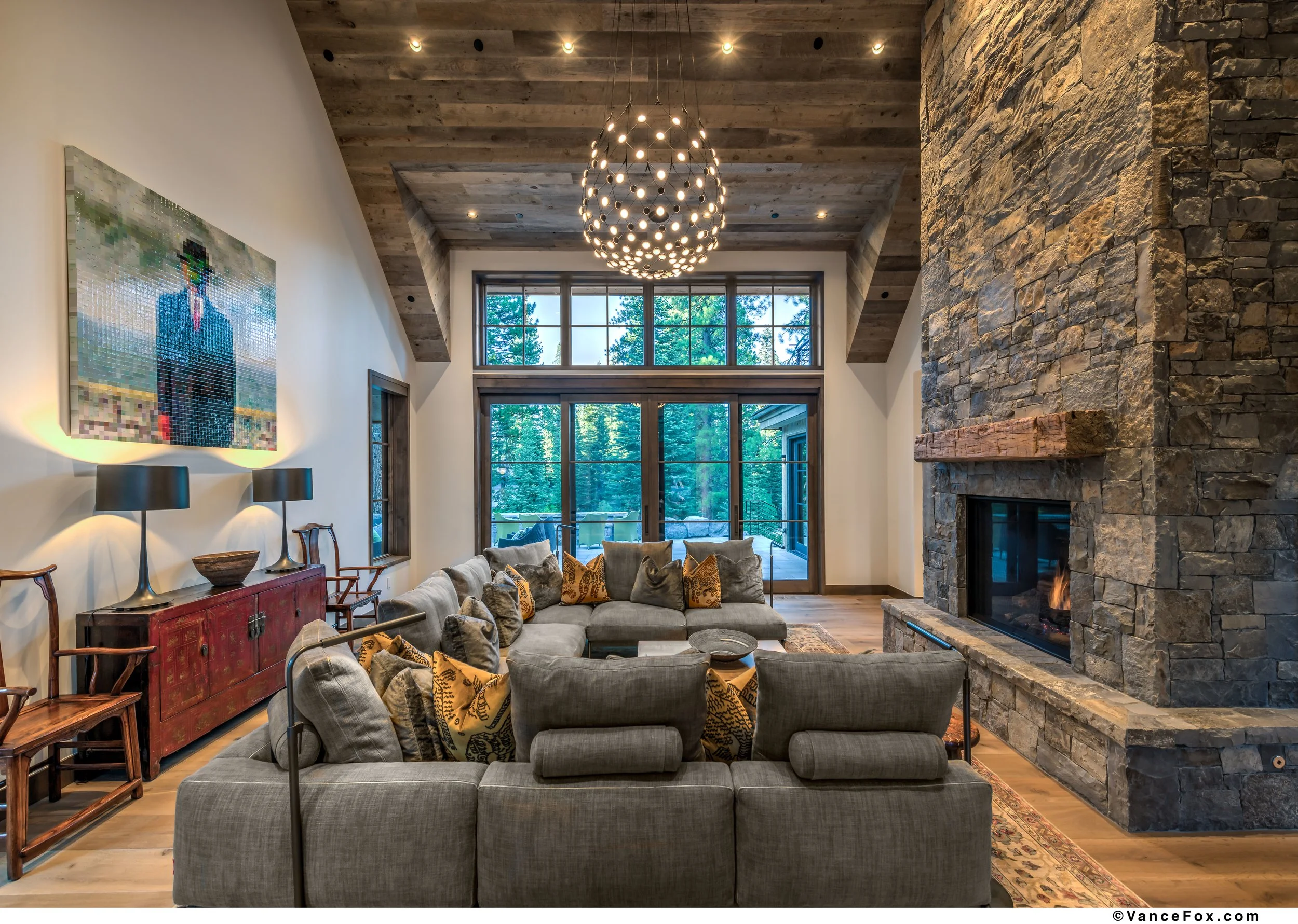 A living room with a large stone fireplace, wooden ceiling, modern chandelier, gray sectional sofa with patterned pillows, and glass doors leading to a yard with trees.