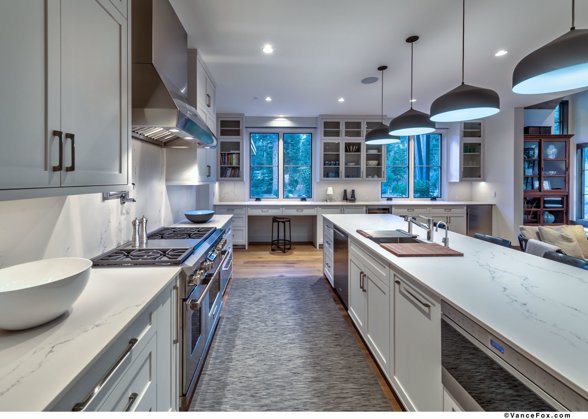 Modern kitchen with white cabinets, marble countertops, and pendant lights, overlooking outdoor scenery through multiple windows.