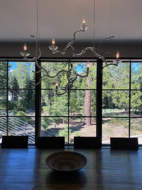 Modern chandelier with multiple branches and candle-shaped bulbs hanging above a dining table in front of large window with trees outside.