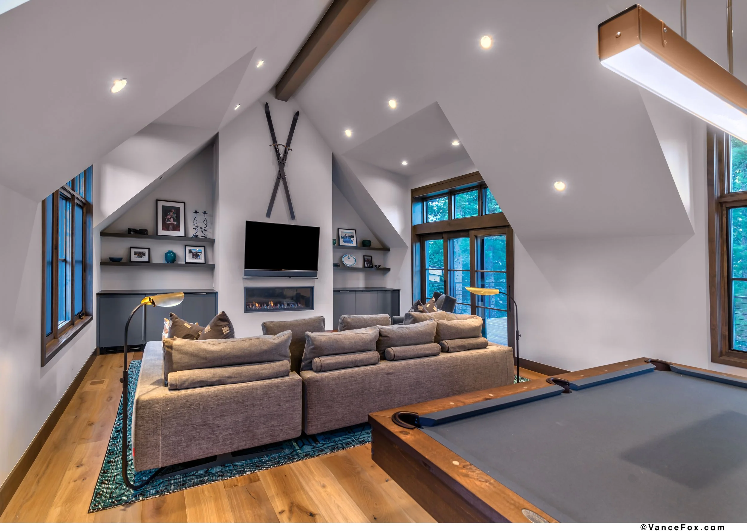 Living room with sloped ceiling, gray sofa, pool table, and large windows with trees outside.