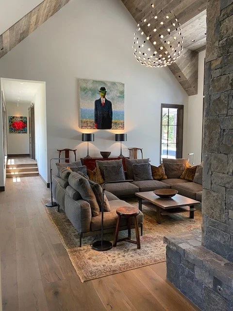 Living room with a vaulted ceiling, stone fireplace, gray sectional sofa, wooden coffee table, and artwork of a man in a suit with a green face on the wall.