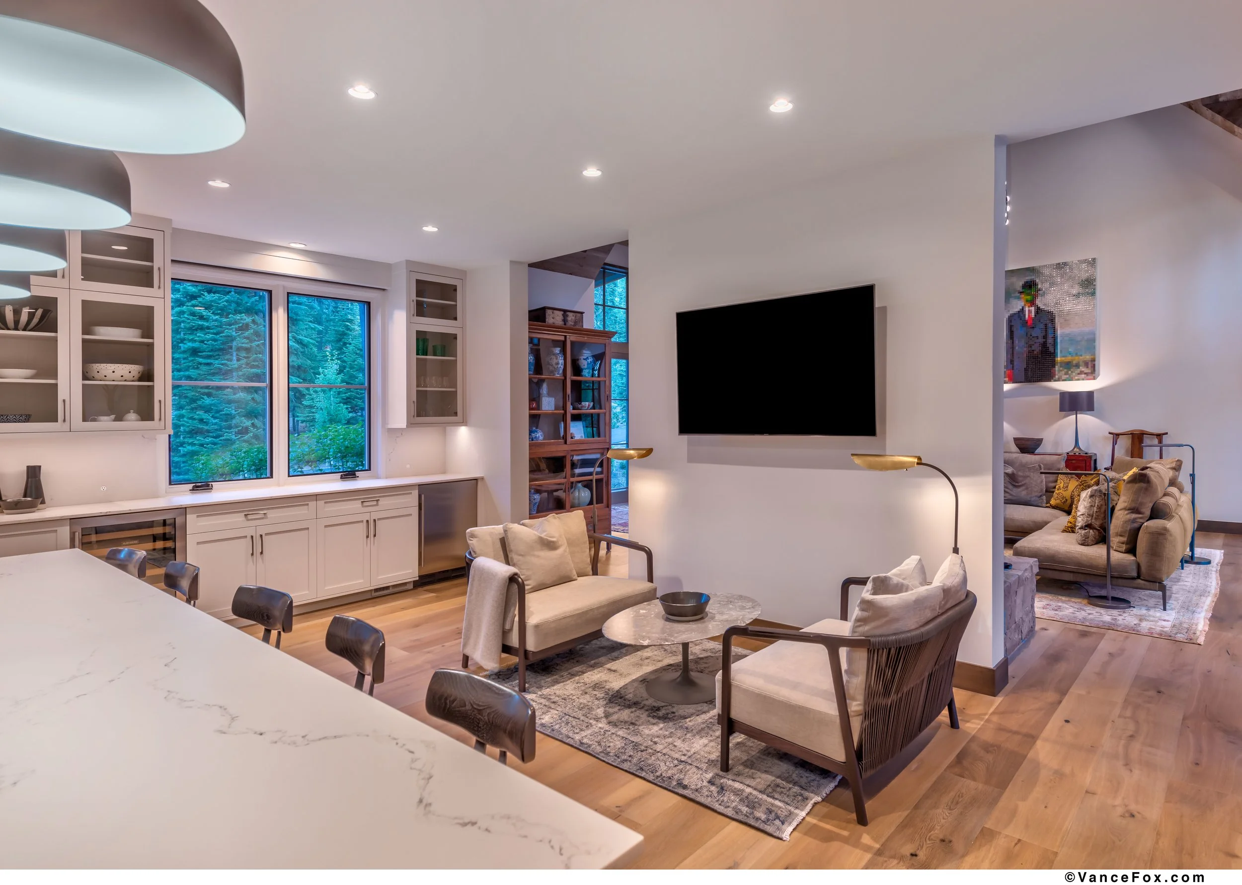 Modern living room with a wall-mounted TV, beige armchairs, a marble coffee table, wooden flooring, and an open kitchen with white cabinets and a window showing trees outside.