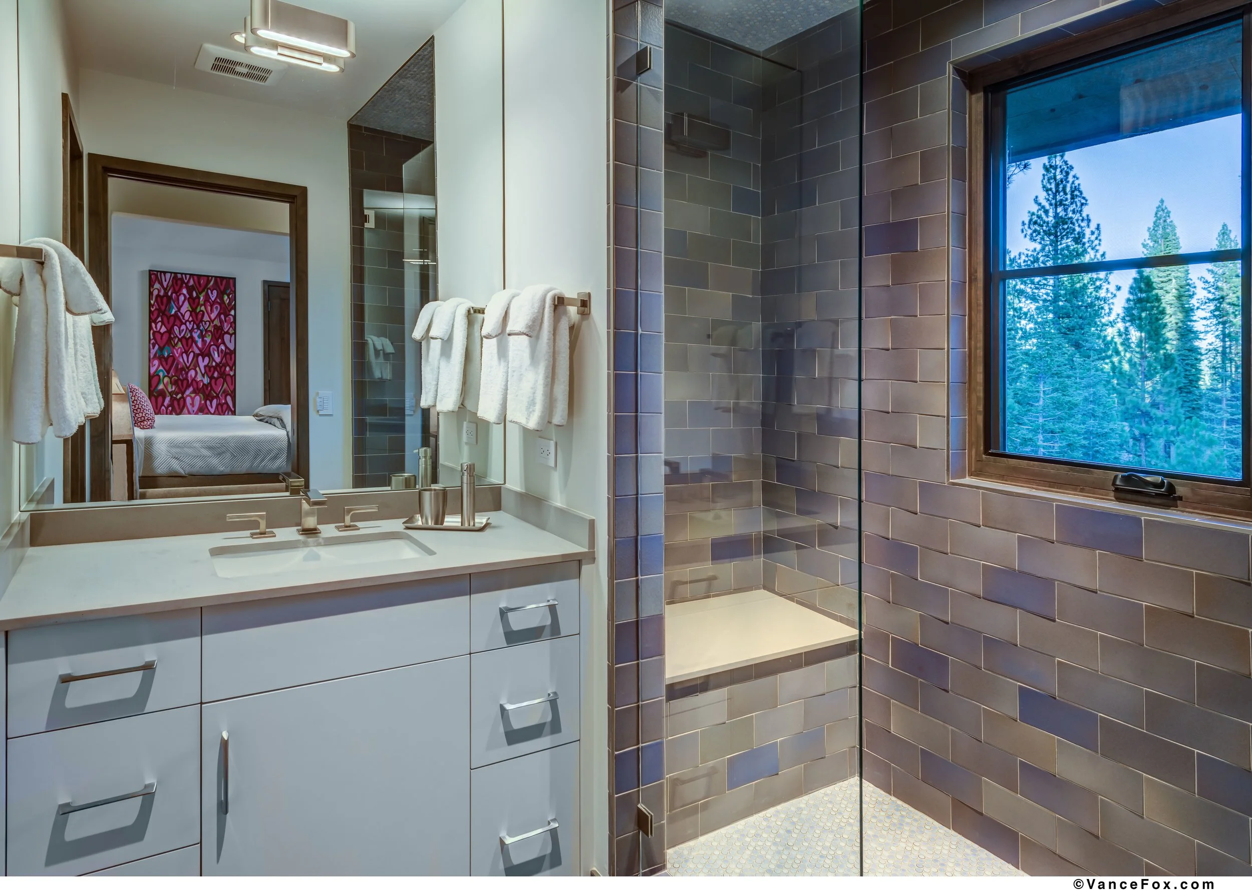 Modern bathroom with white vanity, large mirror, towels, glass-enclosed shower with dark tiles, and a window showing trees outside.