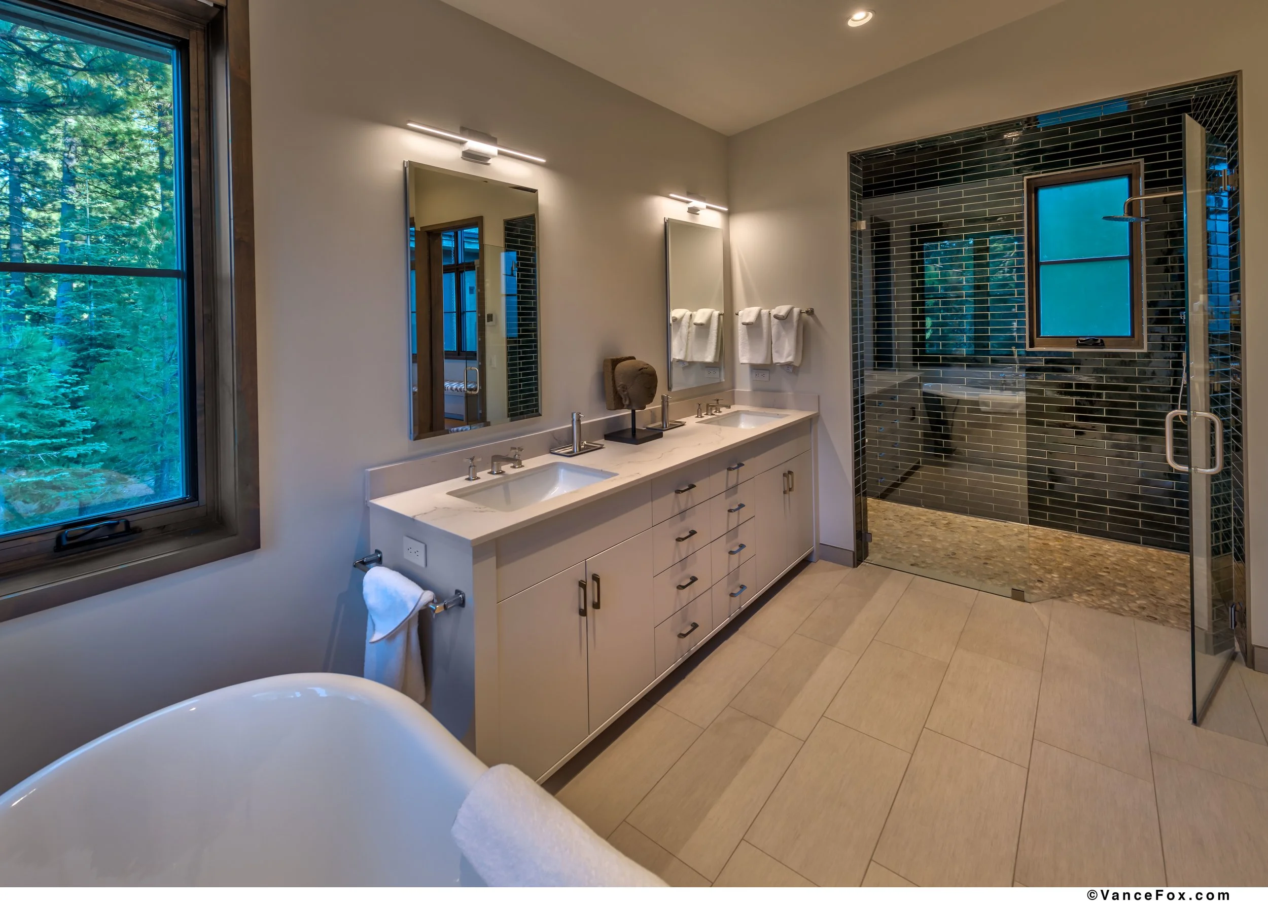 Modern bathroom with a double vanity, large mirror, bathtub, and a walk-in shower with dark tiles and a window.
