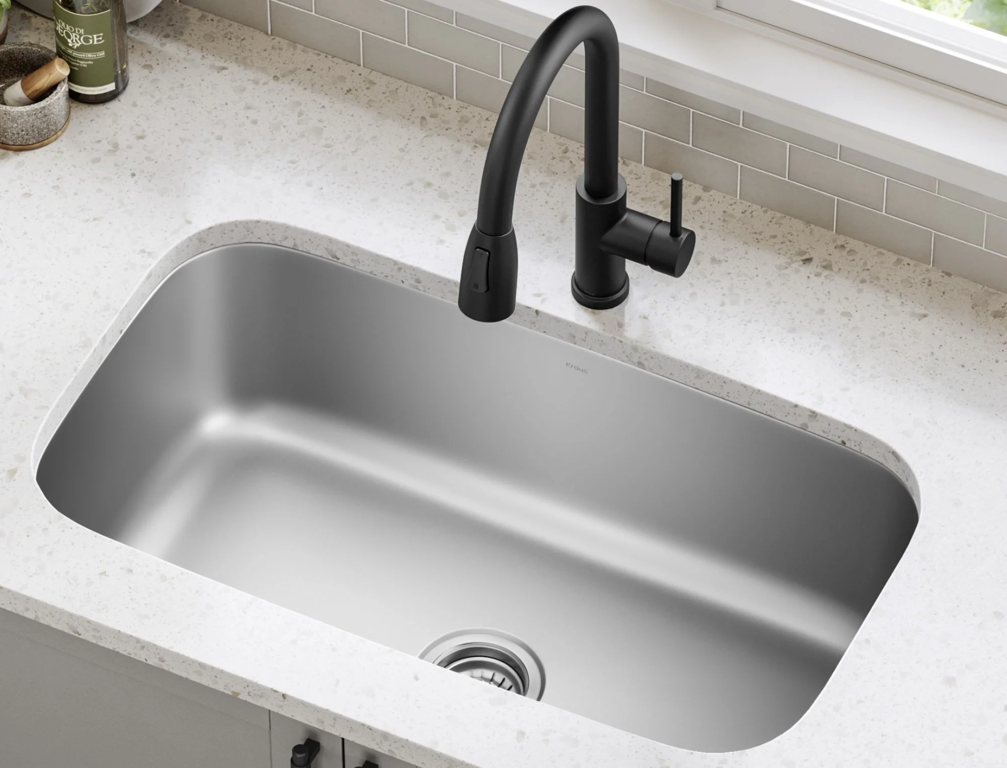 Kitchen sink with a black faucet on a white speckled countertop next to a window, with a small container of spices and a bottle in the corner.