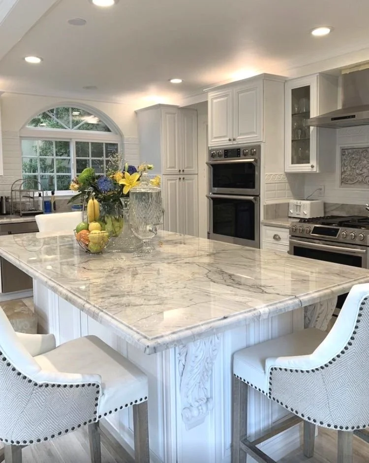 A bright kitchen featuring a marble island with flowers and fruit, white cabinets, stainless steel appliances, and a window with a view of greenery.