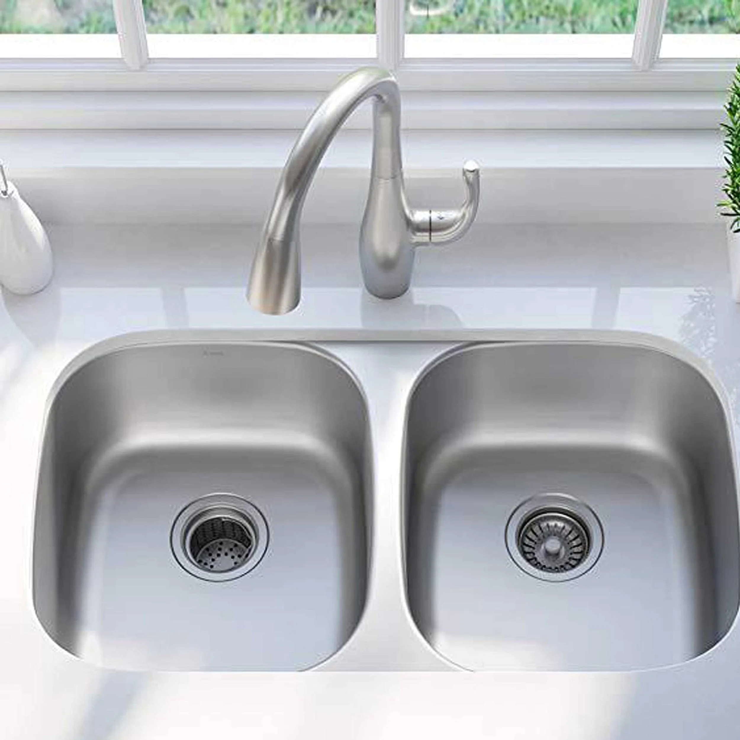 A stainless steel kitchen sink with two basins and a modern curved faucet, set against a white countertop and a window with greenery outside.