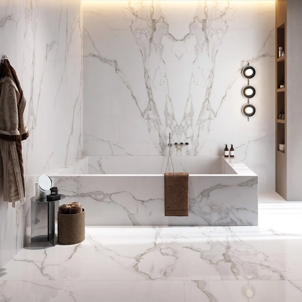 Modern bathroom with white marble walls and flooring, bathtub with a brown towel hanging over its edge, a small side table with bath products, towels in a basket, and a built-in shelf with toiletries. Black circular wall lights are on the right wall.