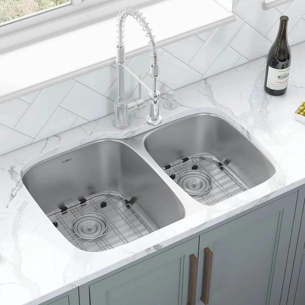 Modern kitchen sink with two basins, stainless steel, with a gooseneck faucet and a drain rack, set in a white marble countertop. A bottle of wine and a bowl of green grapes are on the counter, with a window in the background.