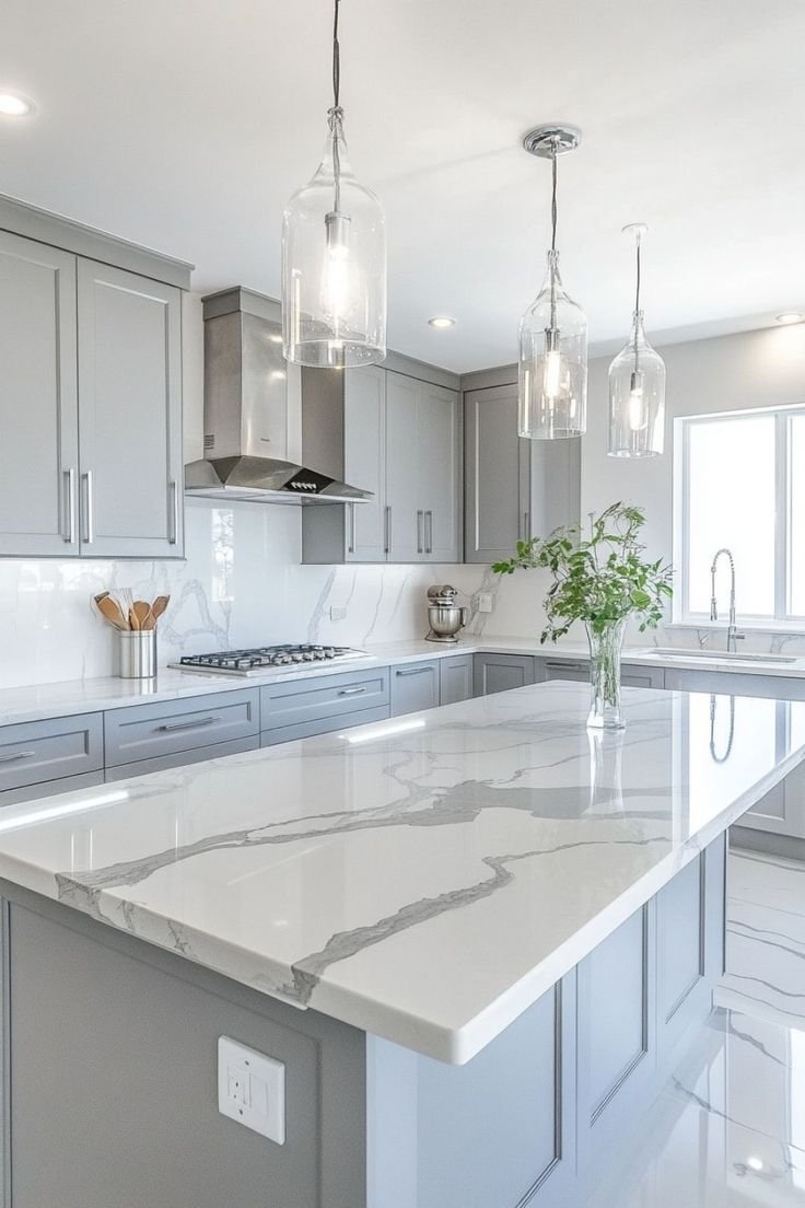 Modern kitchen with gray cabinets, white marble countertops with gray veining, pendant lights, and a vase with green leaves on the island