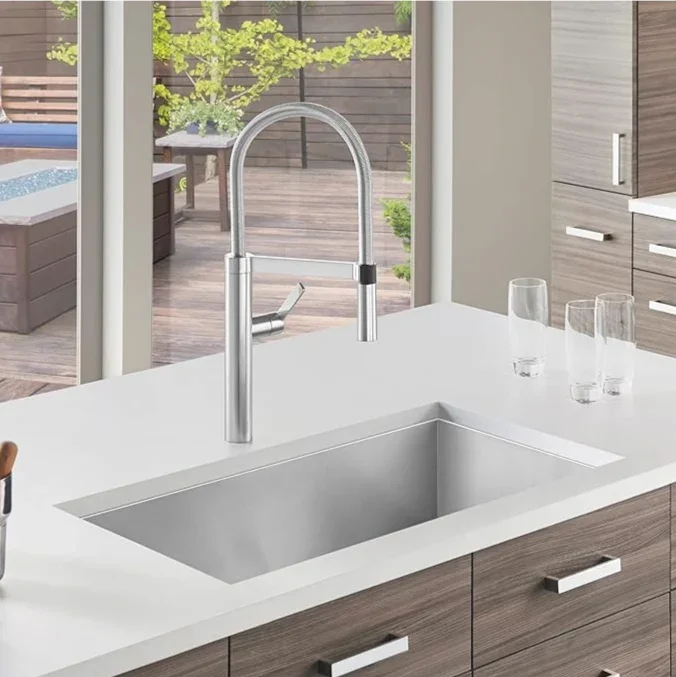 Modern kitchen sink with a high arc chrome faucet and three glasses on a white countertop, overlooking an outdoor patio with a hot tub and greenery.