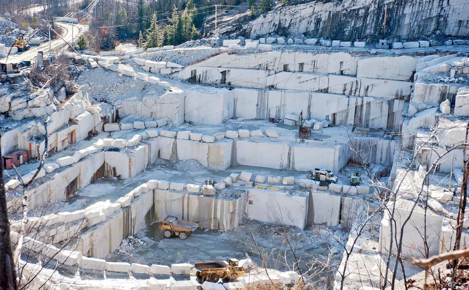 Large limestone quarry with multiple terraced levels and heavy machinery, surrounded by snow and trees.