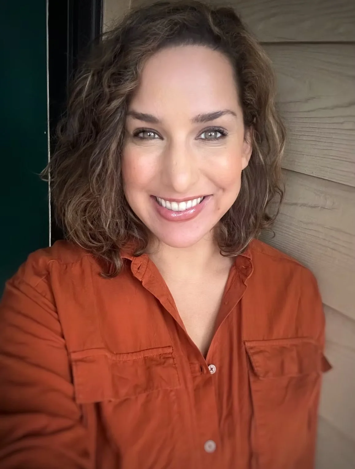 A woman with curly brown hair smiling, wearing an orange shirt, standing next to a wooden wall.