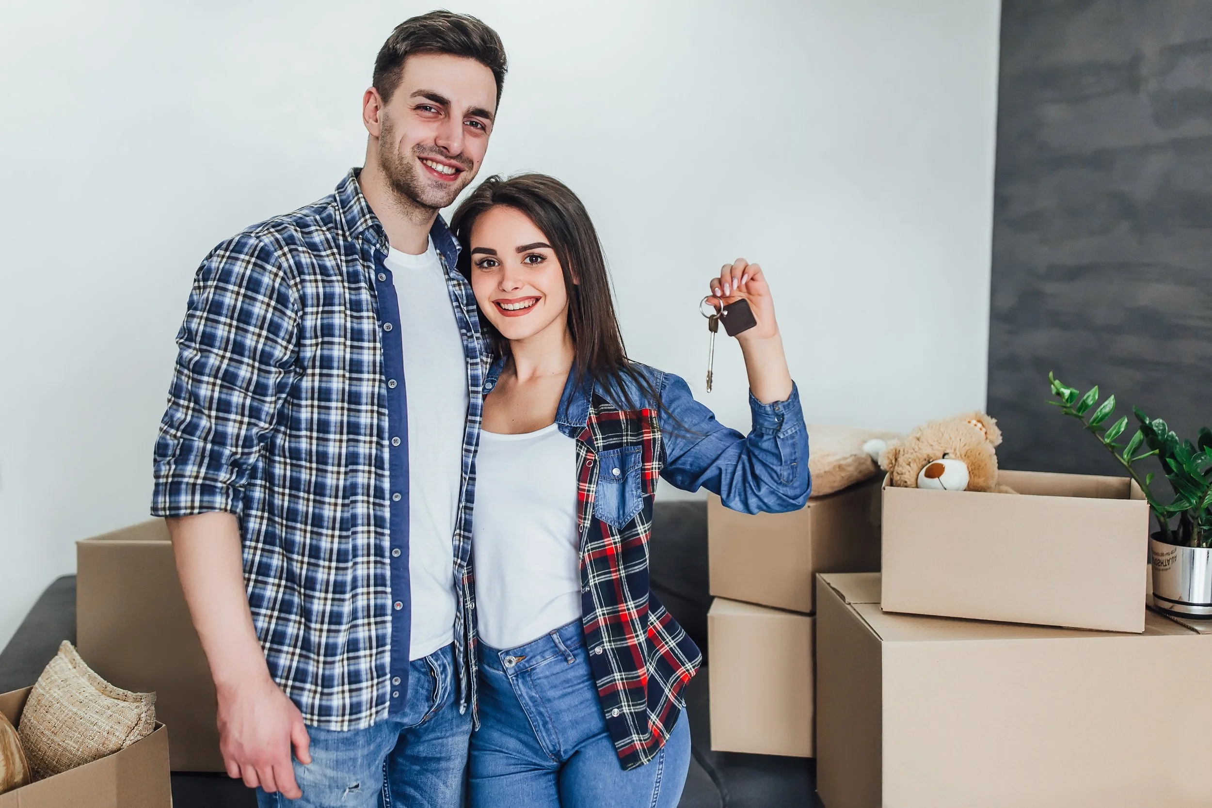 A young couple standing in a room filled with moving boxes, holding a set of keys, smiling at the camera, indicating they are moving into a new home.