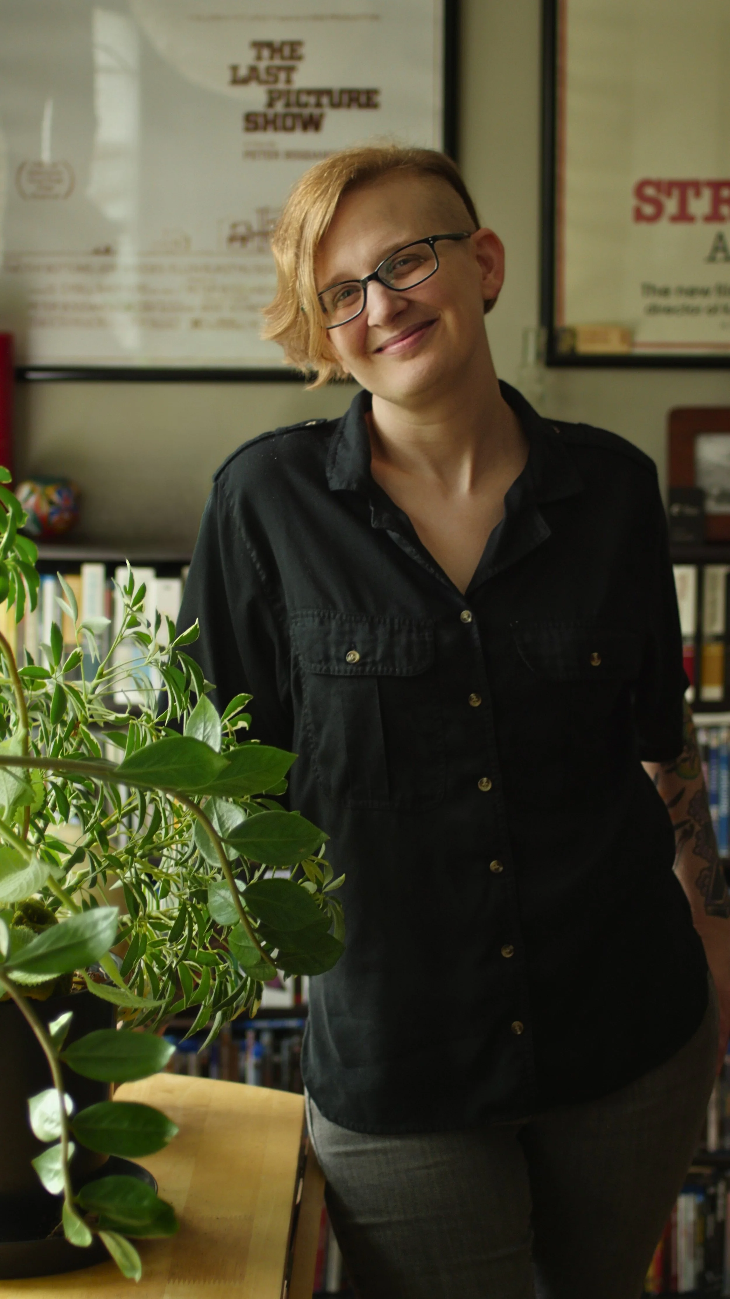 A woman with short blonde hair and glasses, wearing a black shirt, smiling indoors with books and framed posters in the background.