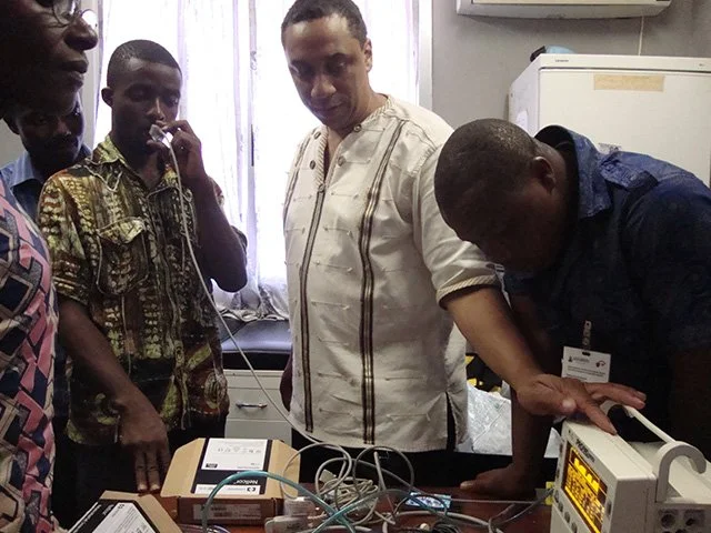 A group of five men gathered around medical equipment in a room, with one man operating a device connected with various wires.