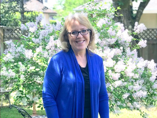 A woman with glasses and blonde hair, smiling, standing outdoors in front of blooming lilac bushes, wearing a blue jacket and black shirt.