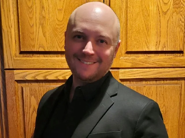 Smiling bald man in black suit standing in front of wooden cabinets.