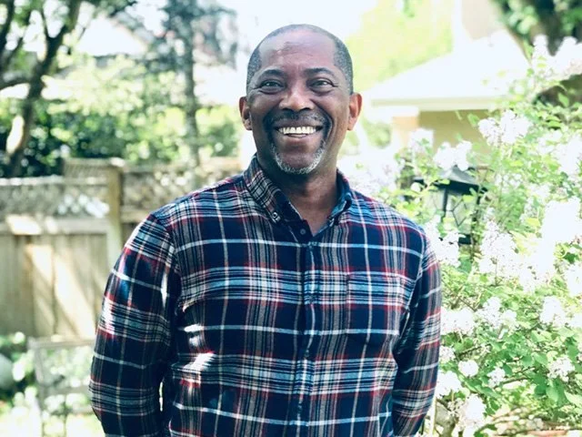 Smiling man standing outdoors in a garden with green foliage and a wooden fence in the background.