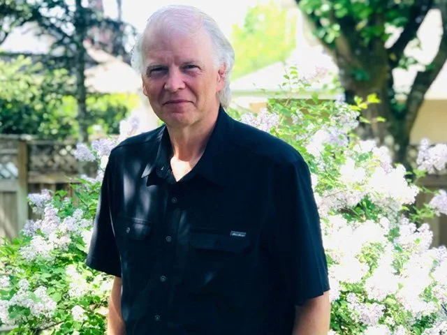 An older man with white hair standing outdoors in a garden with blooming purple and white flowers and green trees in the background.