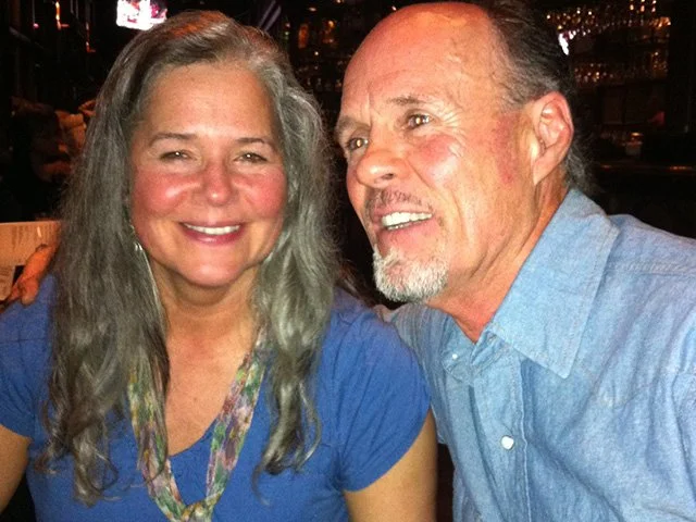A woman and a man smiling in a close-up photo, sitting in a dimly lit restaurant or bar, with a woman having long gray hair and a man with short gray hair and a goatee, both wearing blue shirts.