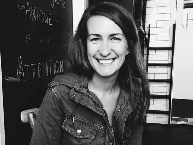 Black and white photo of a woman smiling, wearing a jacket, sitting indoors with a blackboard and stairs in the background.