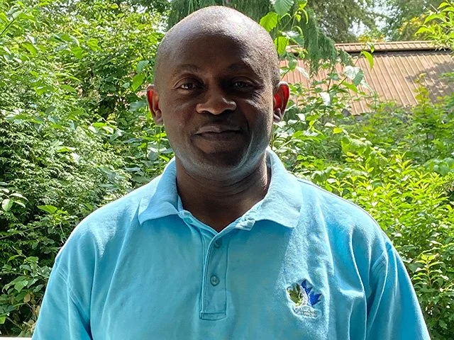 Man in a light blue polo shirt standing outdoors with greenery and a building with a sloped roof in the background.