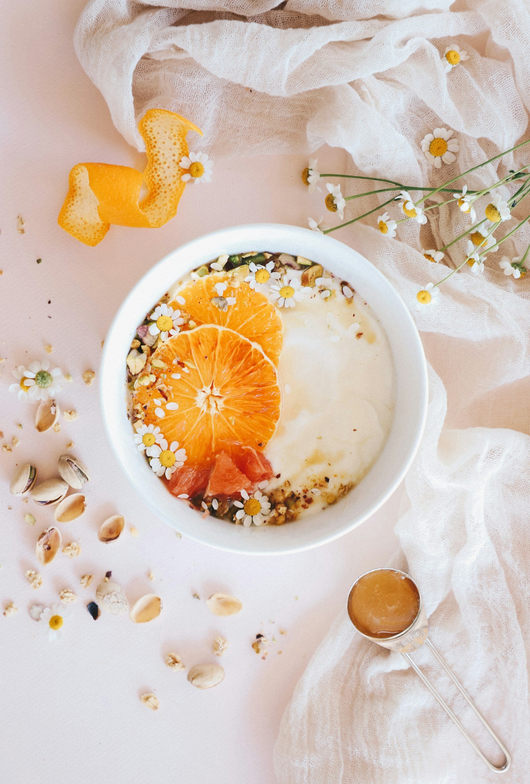 A bowl of yogurt topped with orange slices, mysterious seeds, and white flower petals. Scatterings of pumpkin seeds surround it, with some orange peel and small daisies nearby. A small glass jar containing syrup or honey is at the lower right, and a white gauzy cloth is draped in the background.