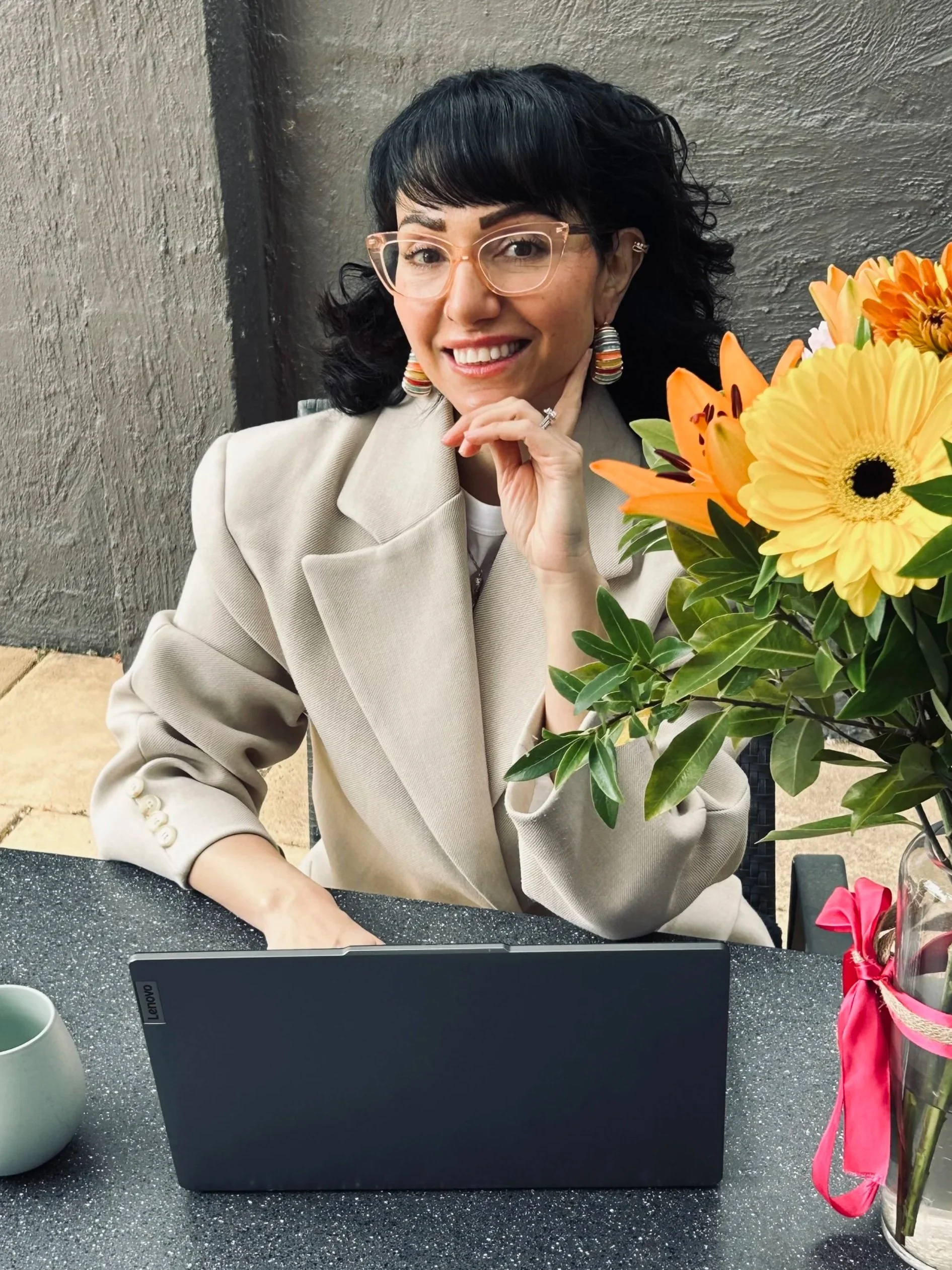 A woman with black wavy hair and glasses smiling, sitting at a table with a bouquet of orange and yellow flowers and a black laptop in front of her.