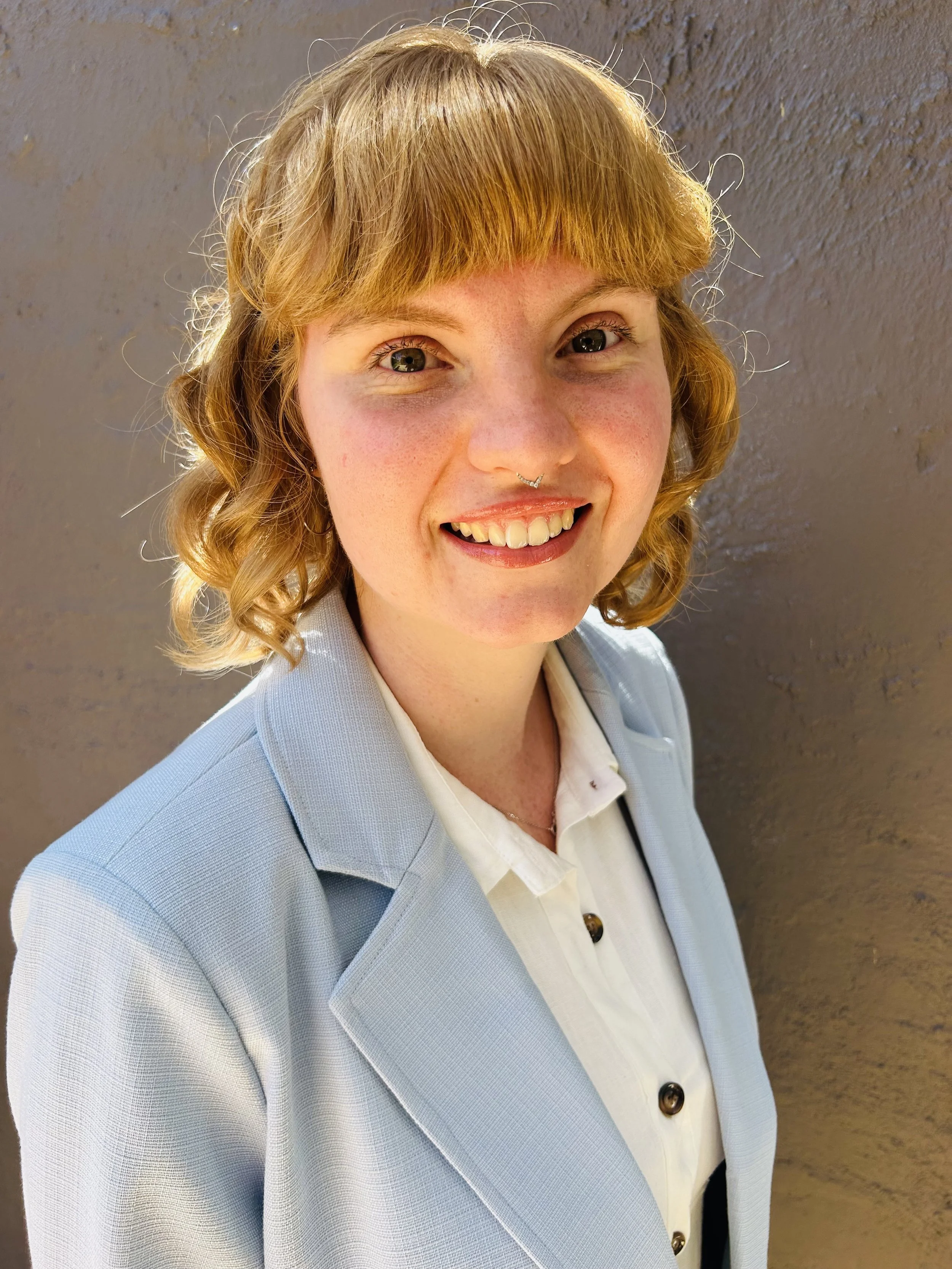Close-up of a woman with curly blonde hair smiling, wearing a pale blue blazer over a white shirt, standing against a textured beige wall.