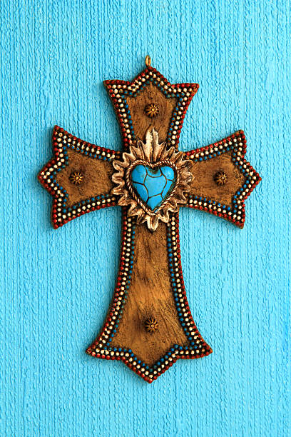 Wooden cross decorated with beads and a blue heart-shaped stone, hanging on a textured blue wall.