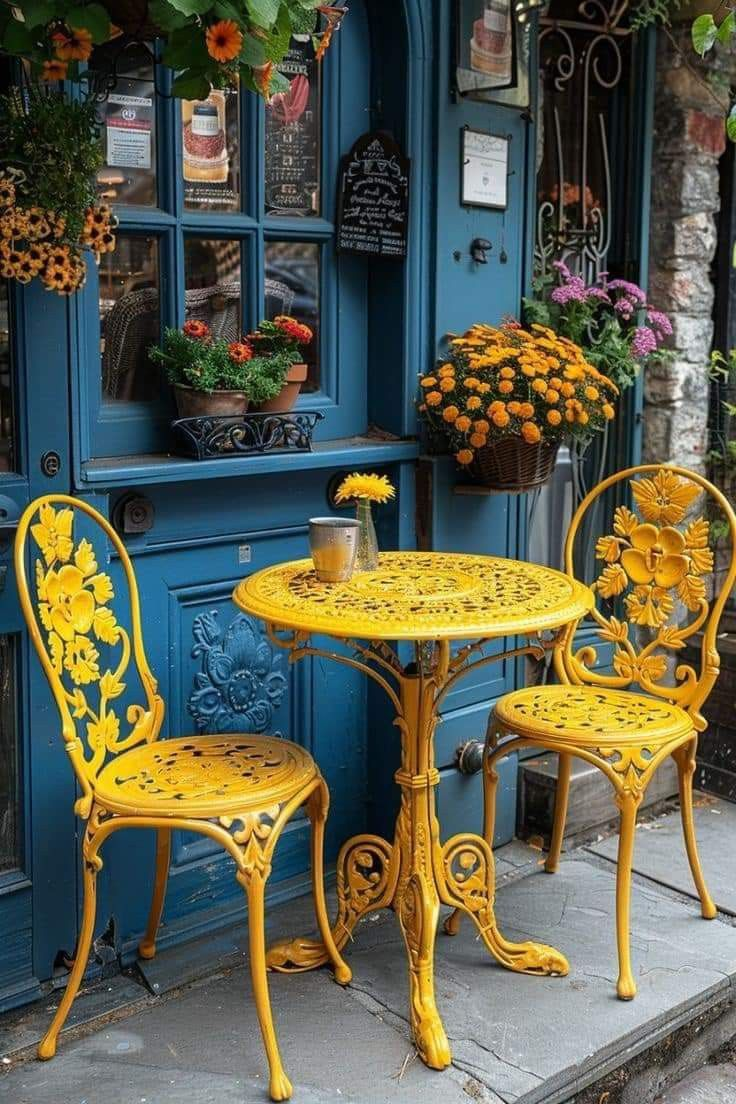 Yellow ornate metal table with two matching chairs outside a blue storefront decorated with potted flowers and hanging baskets.