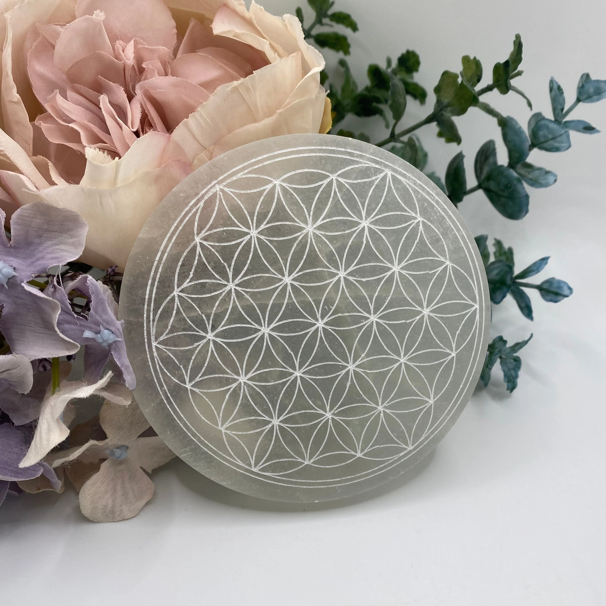 Spherical object with engraved geometric pattern resembling the Flower of Life, placed in front of pastel-colored flowers and greenery.
