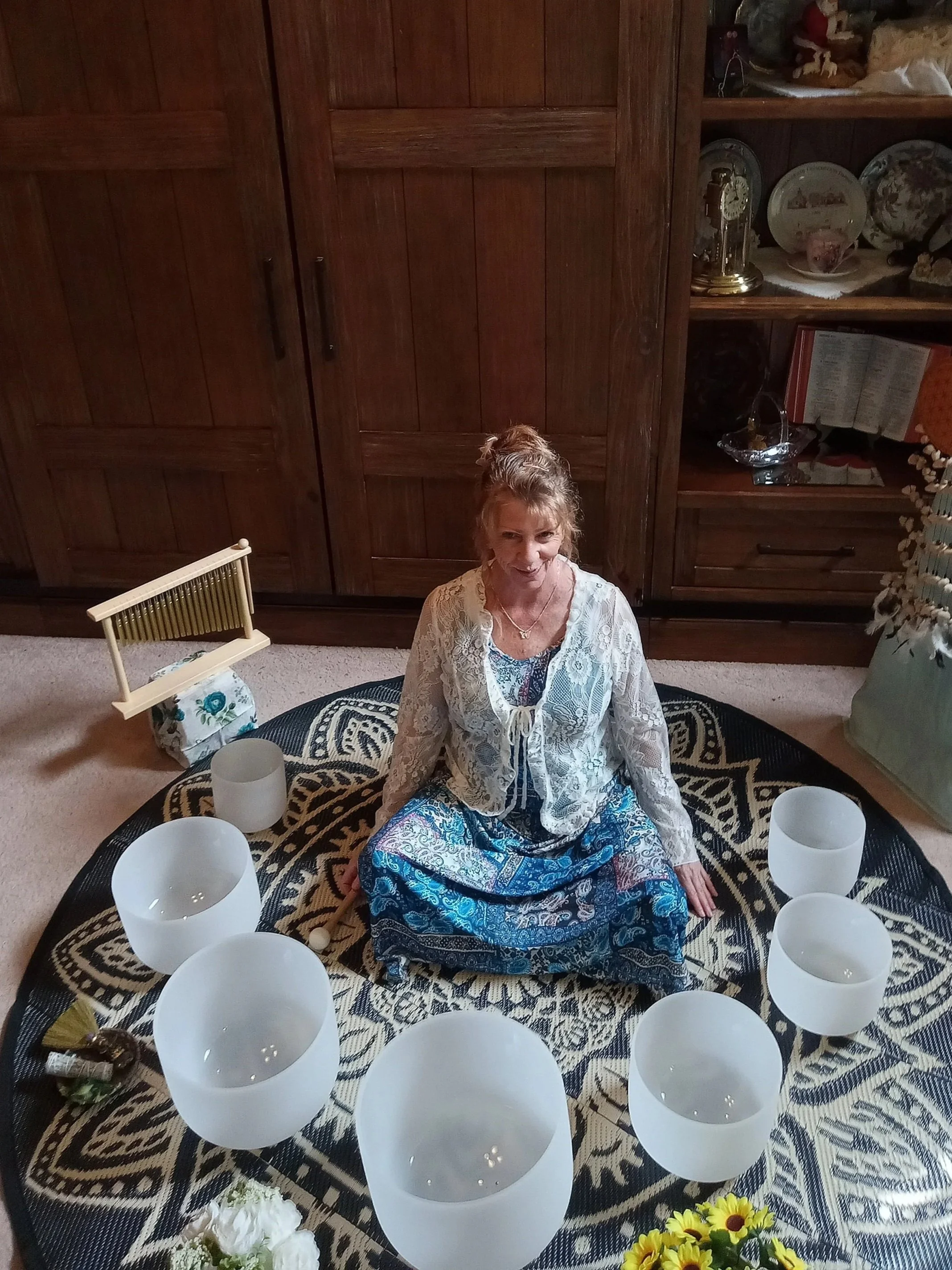 A woman sitting on a decorative rug surrounded by seven glass singing bowls in a room with wooden cabinets.