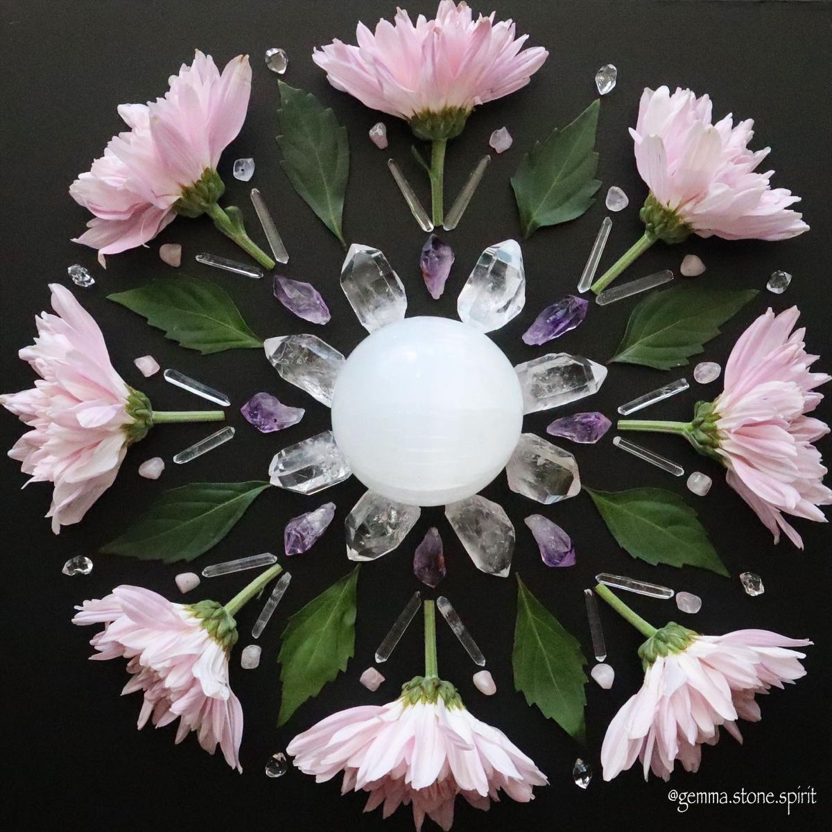 Arrange of pink and white rocks, small crystal points, and sprigs of purple and white flowers on a wooden surface, forming a symmetrical pattern.