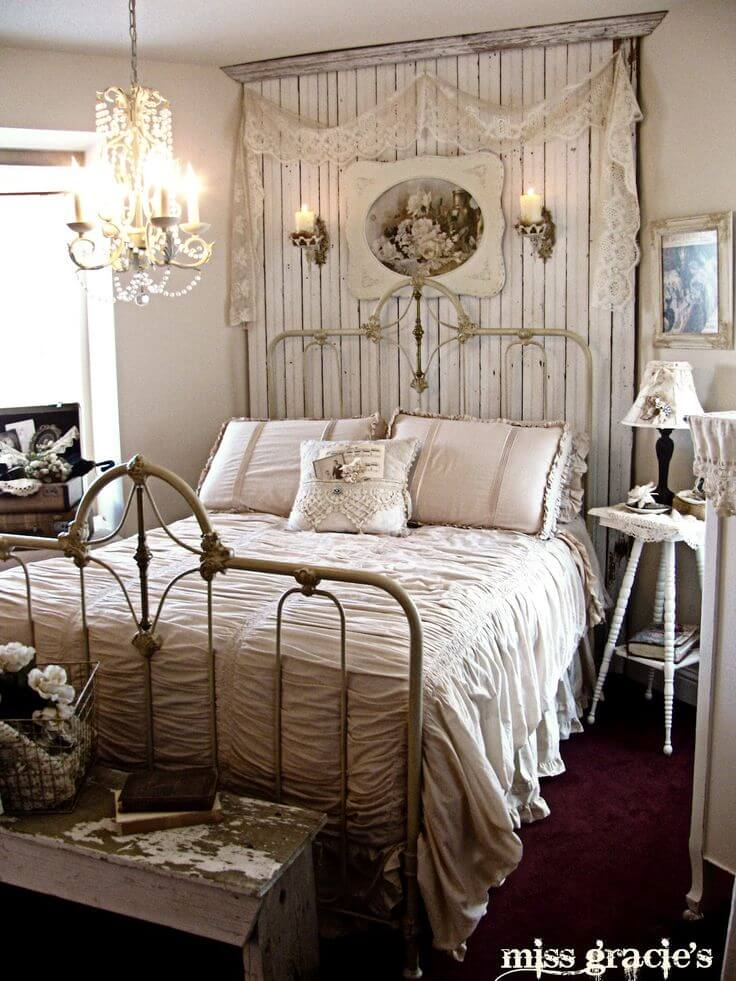 A vintage bedroom with a white metal bed frame, cream-colored bedding, and decorative pillows. An ornate chandelier hangs from the ceiling. Above the bed, there is a wooden wall panel with a floral painting, lace curtains, and candles. A white side table with a lamp and lace cloth is on the right. A small wooden bench with a book and basket of flowers is in front of the bed.