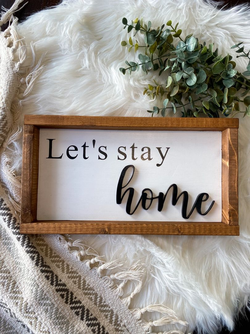 A decorative sign with a wooden frame that says "Let's stay home," placed on a white fluffy rug with a green leafy plant nearby.