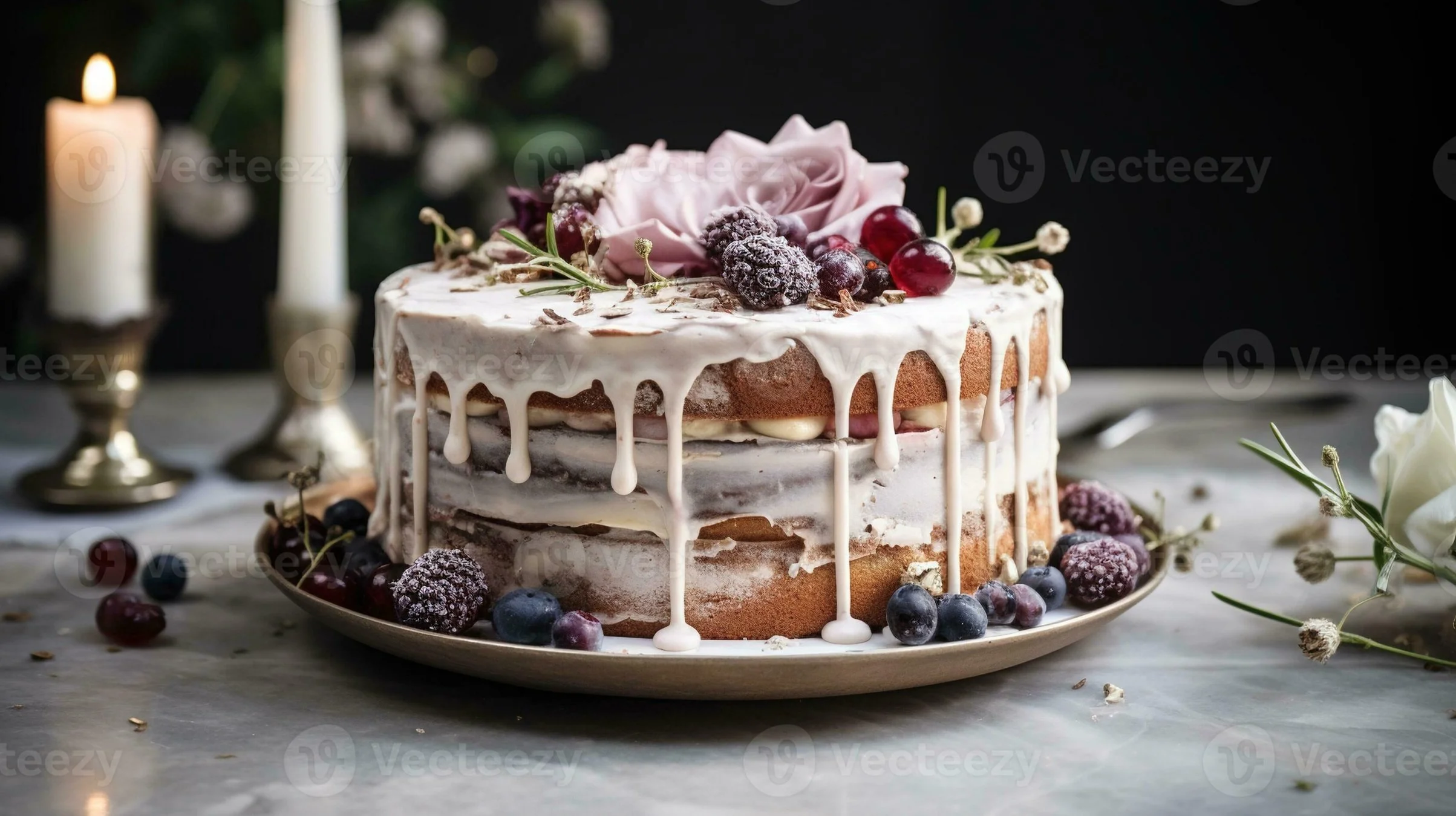 A layered cake with white icing dripping down the sides, decorated with a pink rose, blackberries, red grapes, and edible flowers, placed on a white marble surface with candles and flowers in the background.