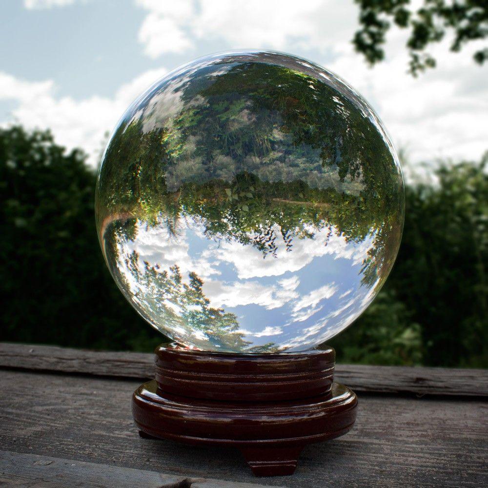 A glass crystal ball reflecting a cloudy sky with trees surrounding it, placed on a wooden surface outdoors.