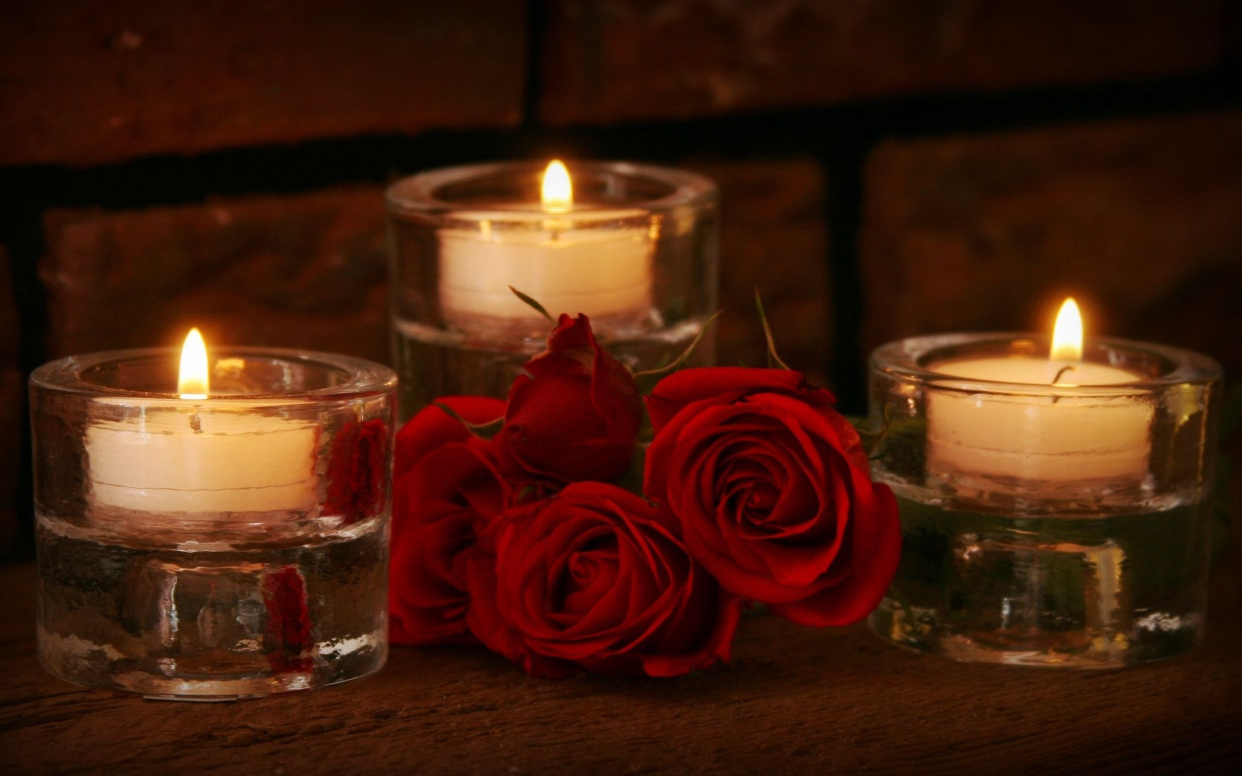 Three lit candles in glass holders with red roses on a wooden surface against a brick wall background.