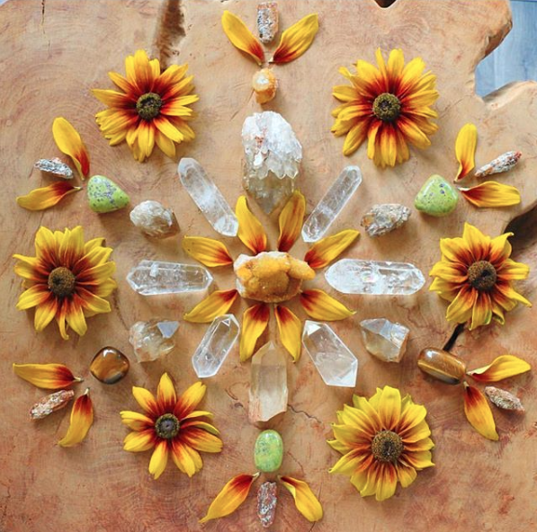 A symmetrical arrangement of yellow and red flowers, green stones, and clear quartz crystals on a wooden surface, resembling a mandala or sacred geometric pattern.